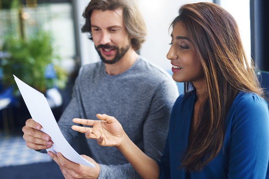 A man and a woman are engaged in discussion while examining a document. They appear to be in a modern indoor setting with plants and a colorful backdrop.