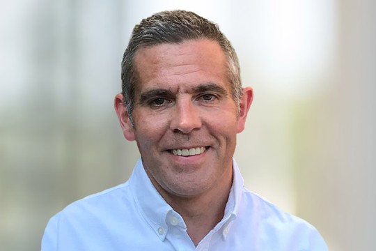 A man with short gray hair smiles, wearing a white collared shirt. He is positioned in a bright, softly blurred outdoor environment, suggesting a casual, friendly atmosphere.
