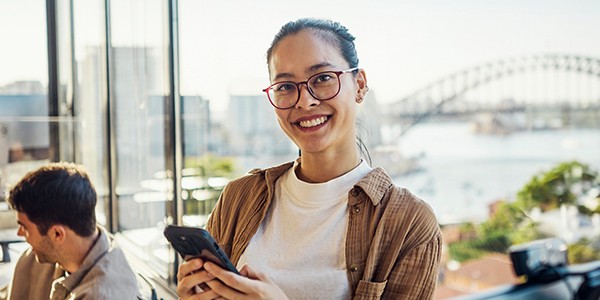 A woman with glasses is smiling and holding a smartphone. She is seated indoors with a glass wall, revealing a view of water and a bridge in the background.
