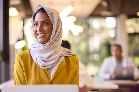 A woman wearing a light-colored hijab and a mustard yellow blouse smiles while seated at a table in a modern office, with other individuals working in the background.