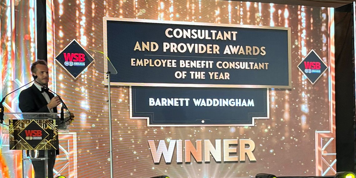 A man in a tuxedo stands at a podium, presenting an award on a stage. The backdrop displays a large sign recognizing "Barnett Waddingham" as the "Employee Benefit Consultant of the Year."