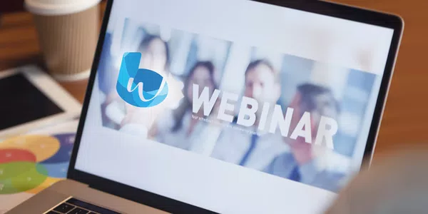 A laptop displays a "WEBINAR" announcement, indicating that a scheduled event will commence shortly. The environment includes a coffee cup and a tablet, suggesting a casual workspace.