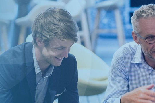 A group of four professionals, including a middle-aged man with glasses, engage in a discussion while seated in a modern, well-lit environment with soft-colored furnishings.