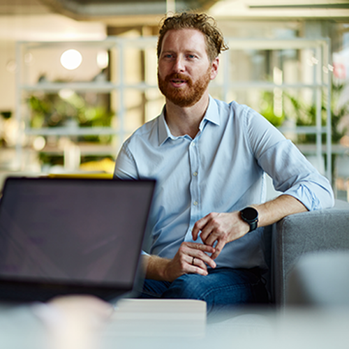A man with a beard sits on a gray sofa, engaging in conversation while holding hands. A laptop is placed in front of him on a table, with an office environment in the background.