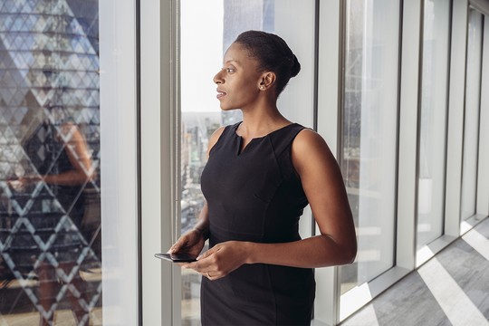 A woman in a black dress stands by large windows, holding a smartphone and looking thoughtfully outside at a city skyline, illuminated by natural light.