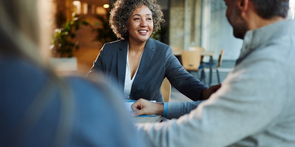 A woman with curly hair is smiling and reaching out to a man during a conversation at a modern office space, which features soft lighting and greenery in the background.