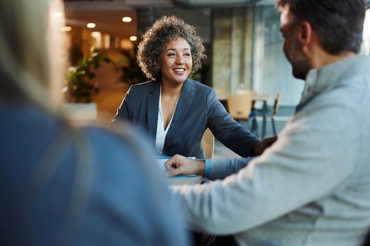 A woman with curly hair is smiling and reaching out to a man during a conversation at a modern office space, which features soft lighting and greenery in the background.