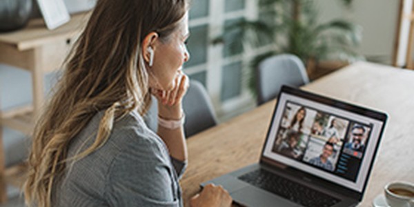 A woman with long hair sits focused at a laptop on a wooden table, using earbuds while engaging in an online video call within a cozy, well-lit room adorned with plants.