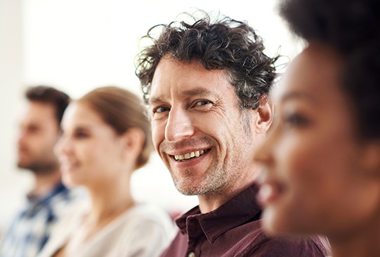 A smiling man sits in a row with three others, engaged in a conversation or event in a bright indoor setting, conveying a sense of friendliness and attentiveness.