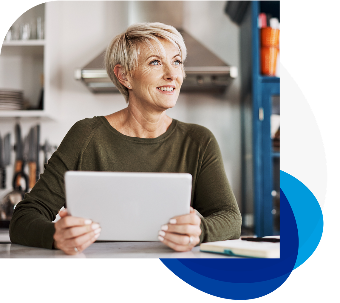 A woman with short hair sits at a kitchen counter, smiling while holding a tablet. The kitchen background features shelves with utensils, a stove, and colorful items.