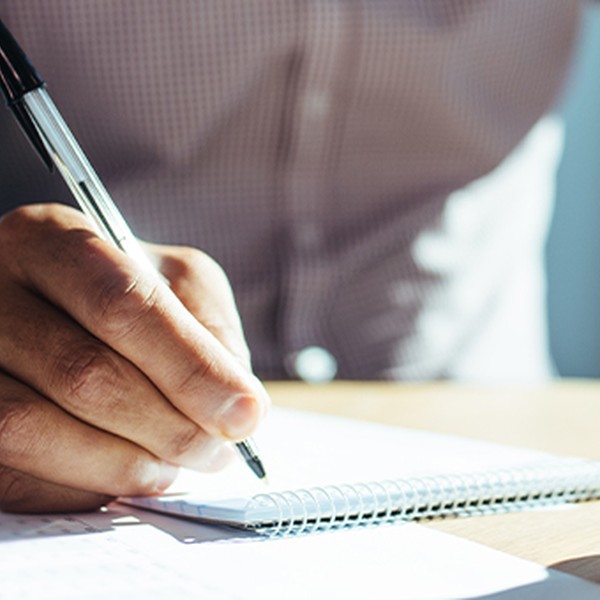 A hand holds a pen, writing on a notepad, while another hand cradles a coffee cup. Surrounding elements include sheets of paper and a smartphone on a wooden table, with a bright, airy background.
