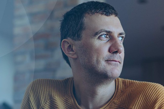 A man with short hair, wearing a textured orange sweater, gazes thoughtfully to the side, set against a blurred indoor background with soft lighting and a stone wall.