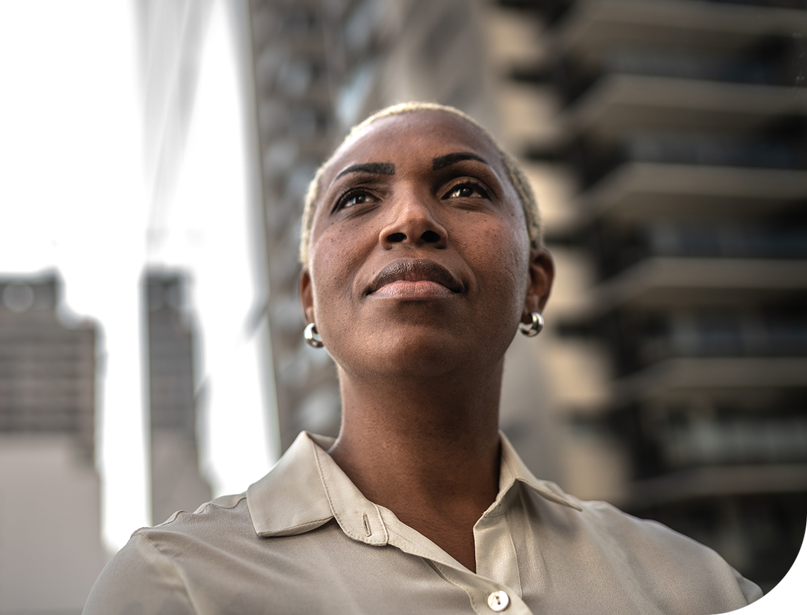 A woman with short, light-colored hair gazes upward confidently. She wears a light shirt and hoop earrings. Modern buildings rise in the background, indicating an urban setting.