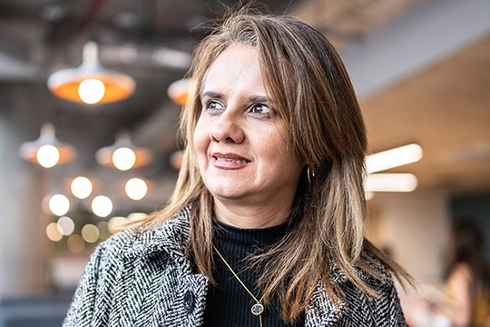 A woman with shoulder-length brown hair smiles softly while looking slightly to the side. She wears a black top and a patterned coat, surrounded by a modern, softly lit environment with blurred lights in the background.