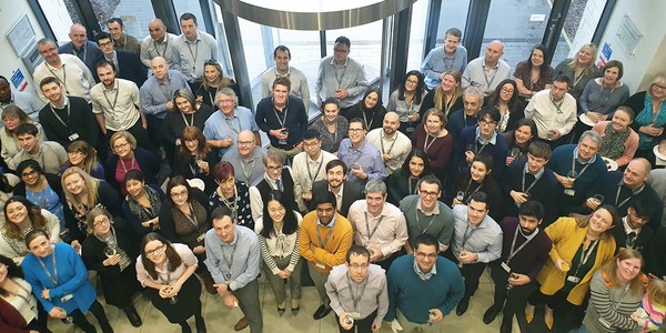 A large group of people, mostly dressed in business attire, are gathered indoors, smiling and holding drinks. They appear engaged and cheerful, likely in a social or celebratory setting.