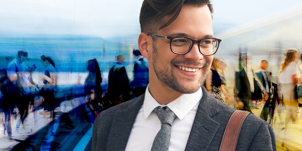 A well-dressed man smiles confidently while standing in a blurred, bustling environment filled with people in formal attire, suggesting a professional or social gathering.