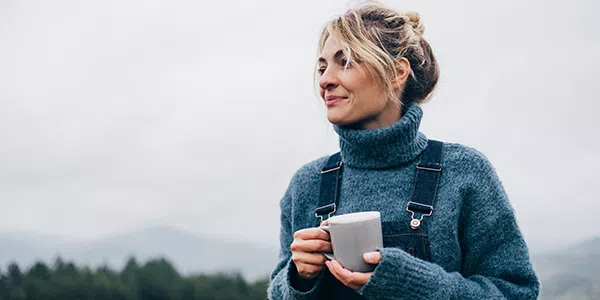 A woman stands outdoors, holding a white mug, wearing a chunky blue sweater and overalls. She gazes thoughtfully into the distance against a backdrop of gray skies and mountains.
