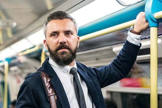 A well-dressed man with a beard stands in a subway car, holding onto a pole for support, appearing contemplative amidst the everyday urban commuting environment.
