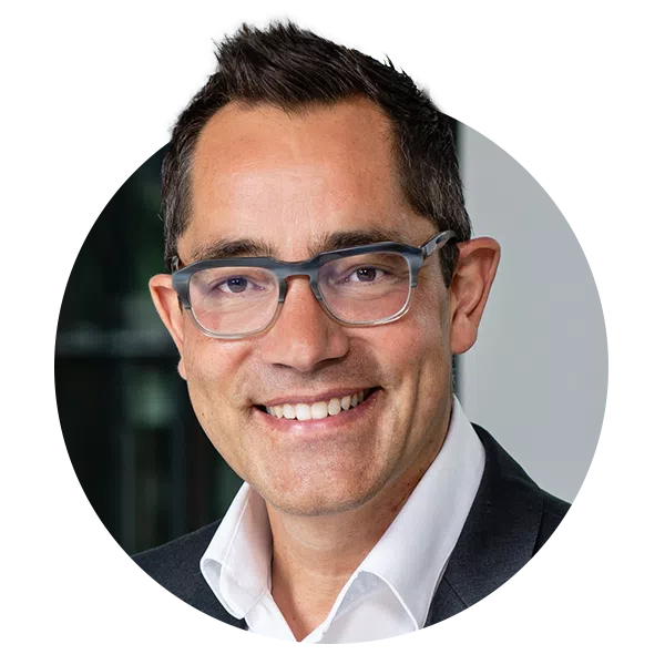 A man with short, dark hair and glasses smiles warmly at the camera. He is wearing a dark suit over a white shirt and is positioned against a blurred background of a modern interior.