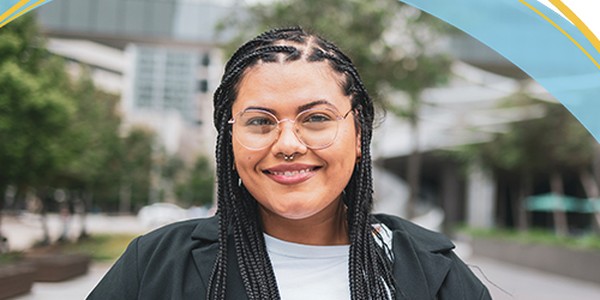 A smiling person with braided hair and glasses stands confidently outdoors. The background features blurred buildings and greenery, suggesting an urban environment. Bright colors arch above the image.