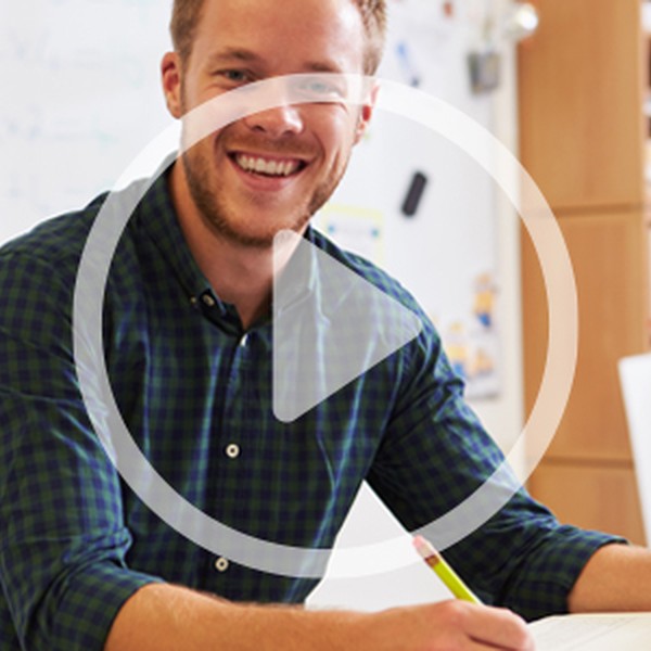 A smiling man wearing a checkered shirt sits at a desk, writing on paper with a pencil. Classroom shelves filled with books are visible in the background, creating a learning environment.