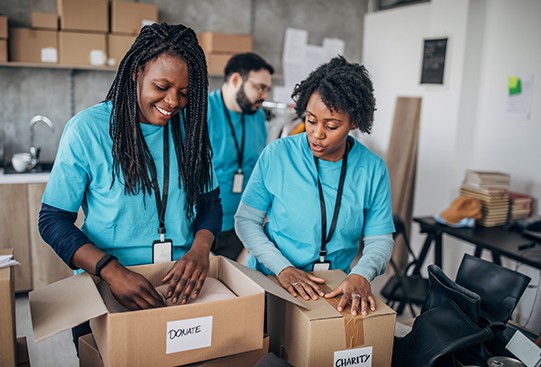 Two people in blue shirts are packing items into labeled boxes marked "DONATE" and "CHARITY." A third person stands in the background, contributing to an organized workspace filled with boxes.