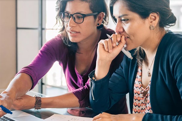 Two women collaborate on a project at a table. One points at a laptop screen while the other looks thoughtfully, holding a pen near her mouth. Bright, modern office setting.