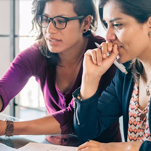 Two women collaborate on a project at a table. One points at a laptop screen while the other looks thoughtfully, holding a pen near her mouth. Bright, modern office setting.