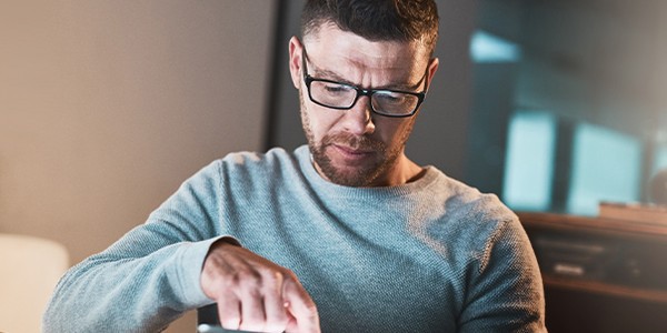 A man in a light sweater is intently using a tablet while seated at a table. The background features soft lighting, enhancing a cozy, focused environment.