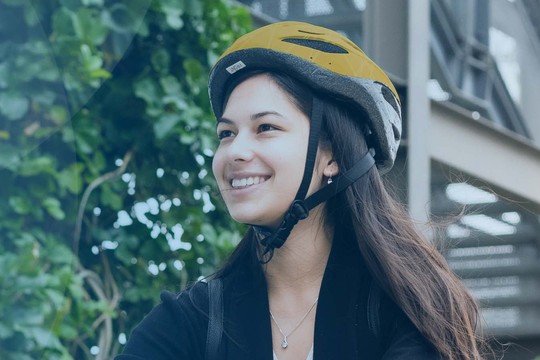 A woman wearing a yellow bicycle helmet smiles while riding a bike. She is surrounded by green foliage and architectural structures, suggesting an urban environment.