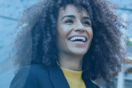A smiling woman with curly hair stands outdoors, wearing a black blazer and yellow top. She exudes joy, with a modern architectural backdrop suggesting a vibrant urban environment.