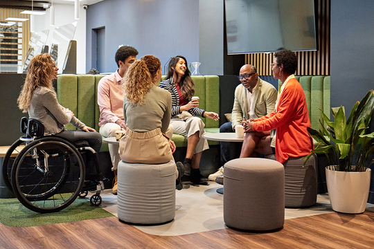 A group of six diverse individuals engage in conversation while seated on round stools around a low table in a modern, relaxed lounge space with greenery and a minimalist design.
