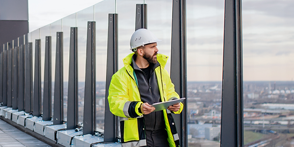 A construction worker in a bright safety jacket and helmet stands near a glass railing on a building's rooftop, holding a tablet and looking out over a cityscape at dusk.