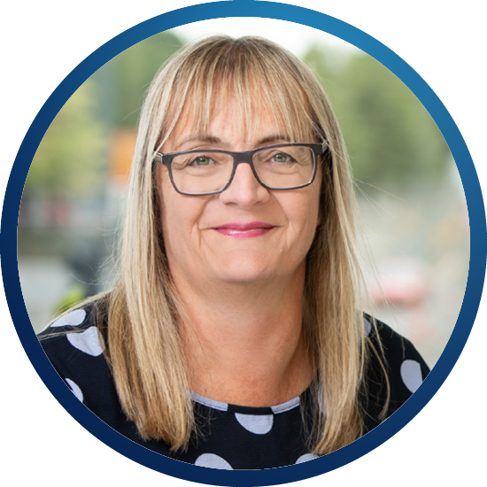 A woman with long, straight hair and glasses smiles warmly, wearing a polka-dotted shirt. The background features an out-of-focus urban scene, suggesting a casual outdoor environment.