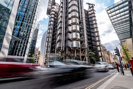 A modern, metallic building with cylindrical structures stands tall amidst glass skyscrapers. Cars speed by on the street, under a partly cloudy sky, creating a dynamic urban scene.