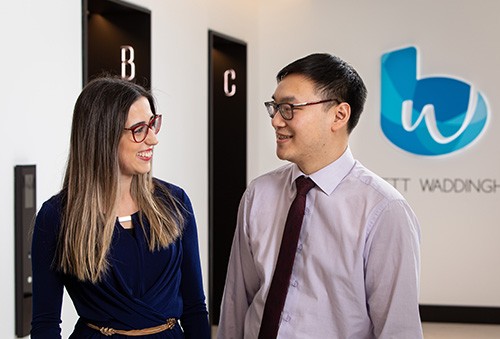 Two professionals, a woman in a blue top and a man in a light purple shirt, are smiling and engaging in conversation. They are indoors, near elevator doors, with a logo in the background.