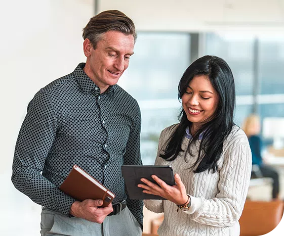 A man and a woman are engaged in conversation, both smiling. The man holds a notebook, while the woman shows him a tablet. They are in a modern office setting with natural light.