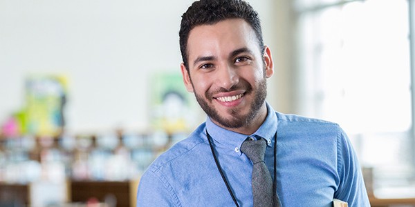 A man in a blue shirt and tie smiles while holding a book. He stands in a bright, spacious room with bookshelves and colorful artwork in the background, suggesting a library or educational setting.