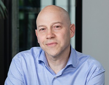 A man with a shaved head and a slight smile sits in an office setting, wearing a light blue shirt. Natural light filters in through nearby windows, creating a warm atmosphere.