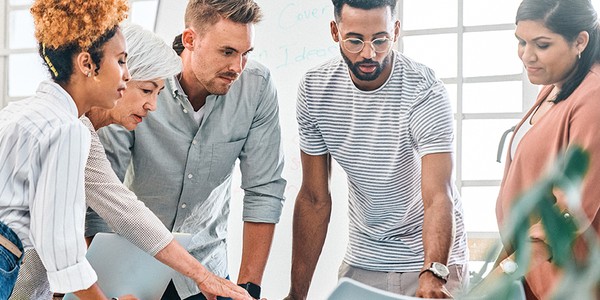 A group of five diverse individuals collaborate around a table, examining documents and digital devices. The setting is a bright, modern office space with large windows.