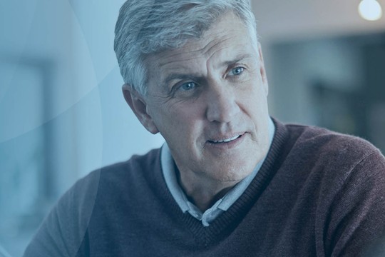 An older man in a sweater engages in conversation, looking attentively. The setting appears bright and modern, with soft colors and blurred background elements suggesting a comfortable indoor space.