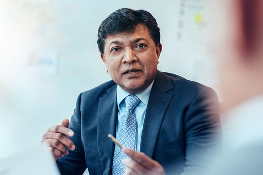 A man in a suit engages in conversation, gesturing with a pen in hand, while seated at a meeting table in a brightly lit office with whiteboard notes in the background.