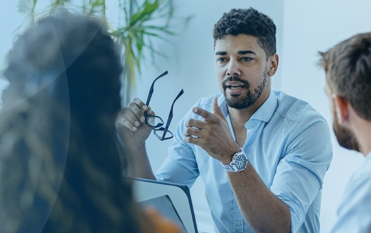 A man gestures while speaking, holding glasses in one hand. He is engaged in a discussion with two other individuals in a bright, modern office with greenery in the background.