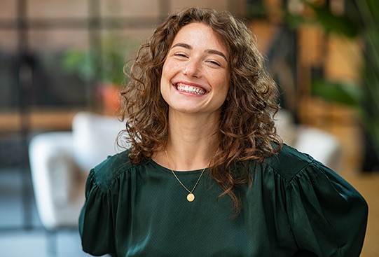 A woman with curly hair smiles brightly, wearing a green top with puffy sleeves. She is in a well-lit, modern indoor environment with plants and soft seating in the background.