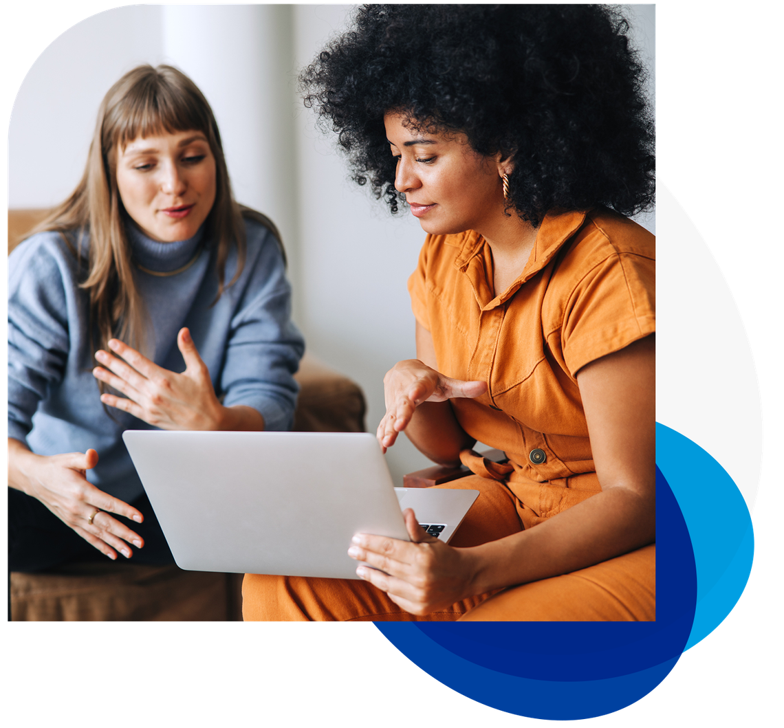 Two women are engaged in conversation, one gesturing animatedly while the other focuses on a laptop in a cozy, modern setting. The first woman wears a blue sweater, and the second is in an orange outfit.