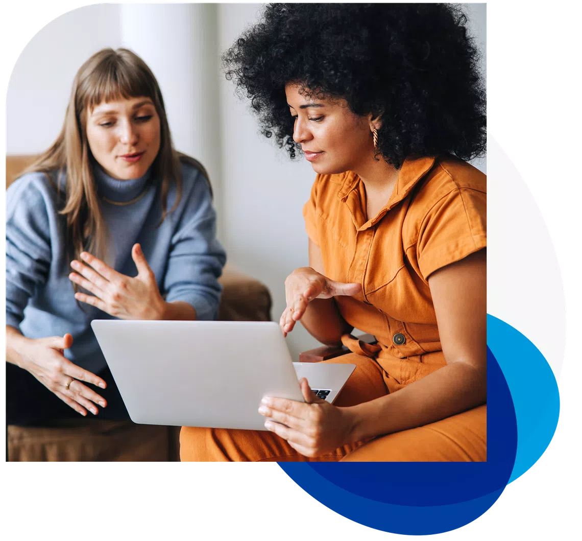 Two women are engaged in conversation, one gesturing animatedly while the other focuses on a laptop in a cozy, modern setting. The first woman wears a blue sweater, and the second is in an orange outfit.