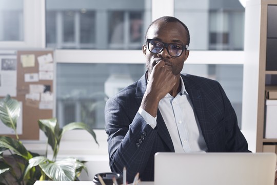 A man in a dark blazer sits at a desk, thoughtfully resting his chin on his hand, surrounded by a plant and a laptop, with a bulletin board in the background.