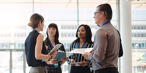 A group of four professionals, holding documents and discussing, stands in a bright, modern office environment with large windows, suggesting a collaborative meeting or brainstorming session.