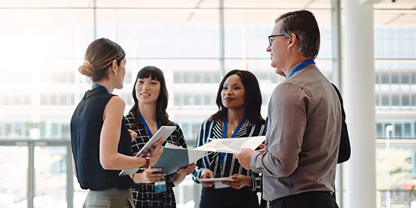 A group of four professionals, holding documents and discussing, stands in a bright, modern office environment with large windows, suggesting a collaborative meeting or brainstorming session.