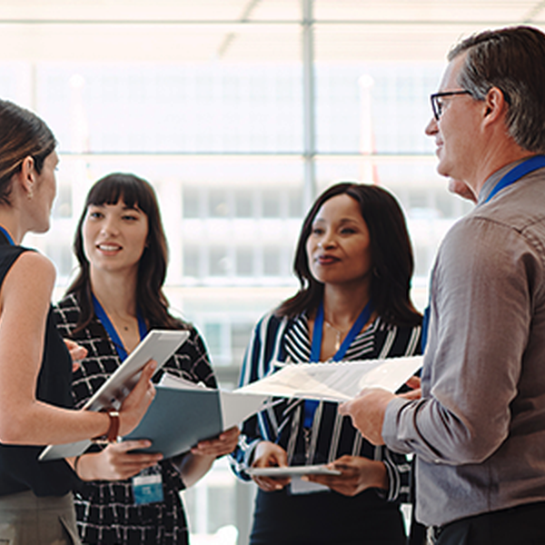 A group of four professionals, holding documents and discussing, stands in a bright, modern office environment with large windows, suggesting a collaborative meeting or brainstorming session.